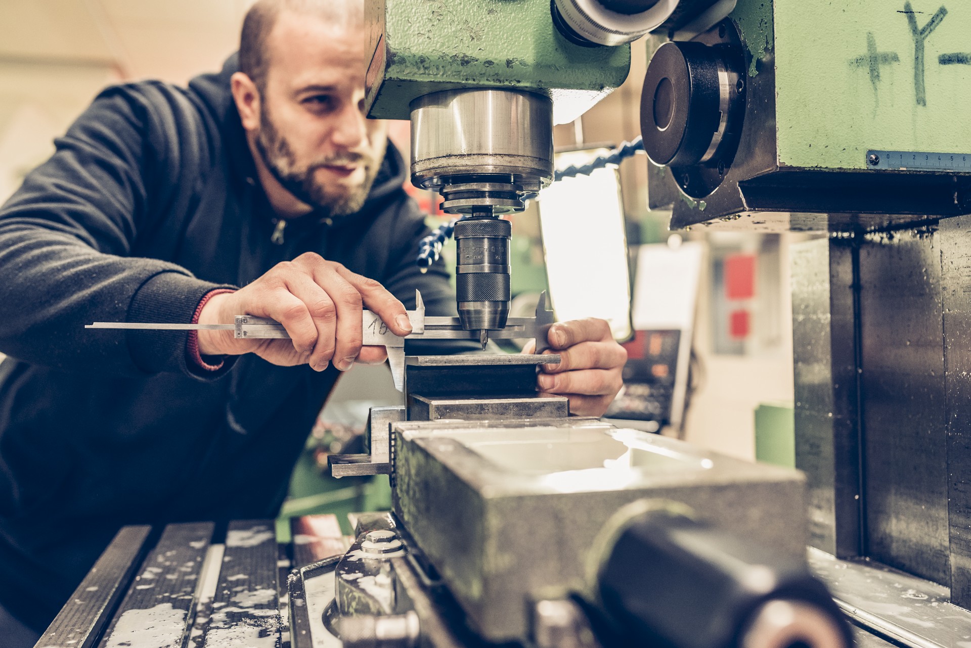 Ein Mann bedient eine Metallfräsmaschine und konzentriert sich intensiv darauf, einen Messschieber an einem Metallstück einzustellen. In der Werkstatt stehen verschiedene Werkzeuge und Maschinen bereit.