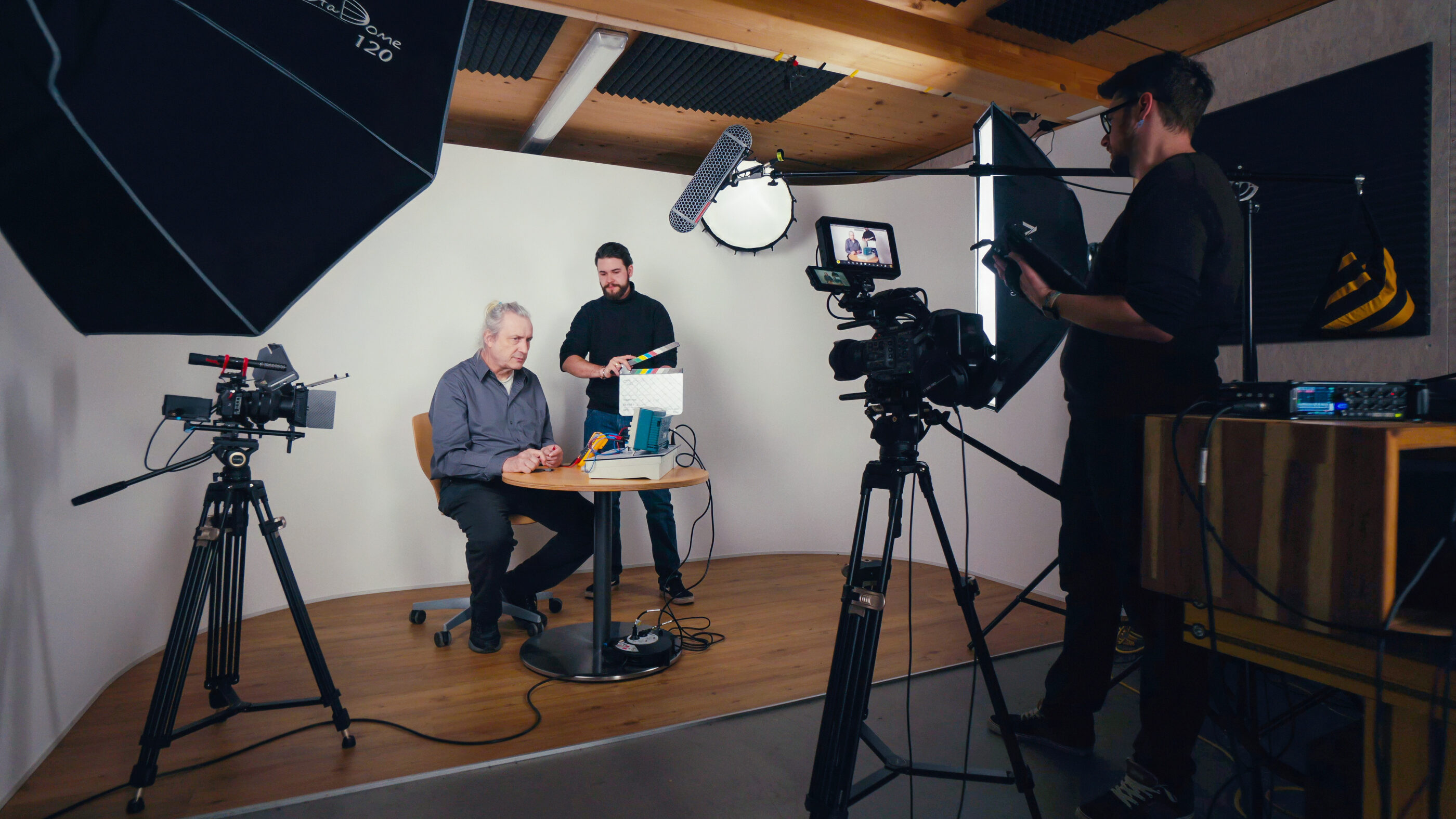Three people are in a video studio. One is sitting at a table with papers and snacks, while another stands holding a clapperboard. Cameras, spotlights, and sound recording equipment surround them, while a third person films.  