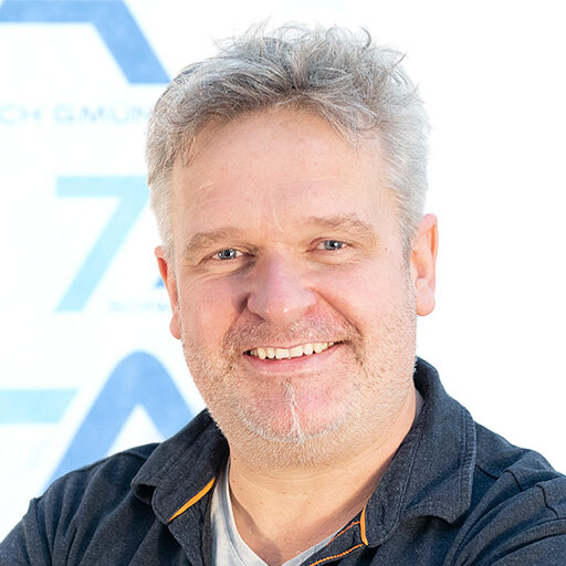 Ein lächelnder Mann mittleren Alters mit hellem Haar und einem kurzen Bart, der ein dunkles Hemd mit Kragen und orangefarbenen Zierstreifen trägt und vor einem weißen Hintergrund mit blauem Text und blauen Formen steht.