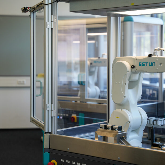 Ein Roboterarm in einem Glasgehäuse arbeitet in einer modernen Labor- oder Fabrikumgebung, in deren Nähe leere Tabletts und Geräte stehen.