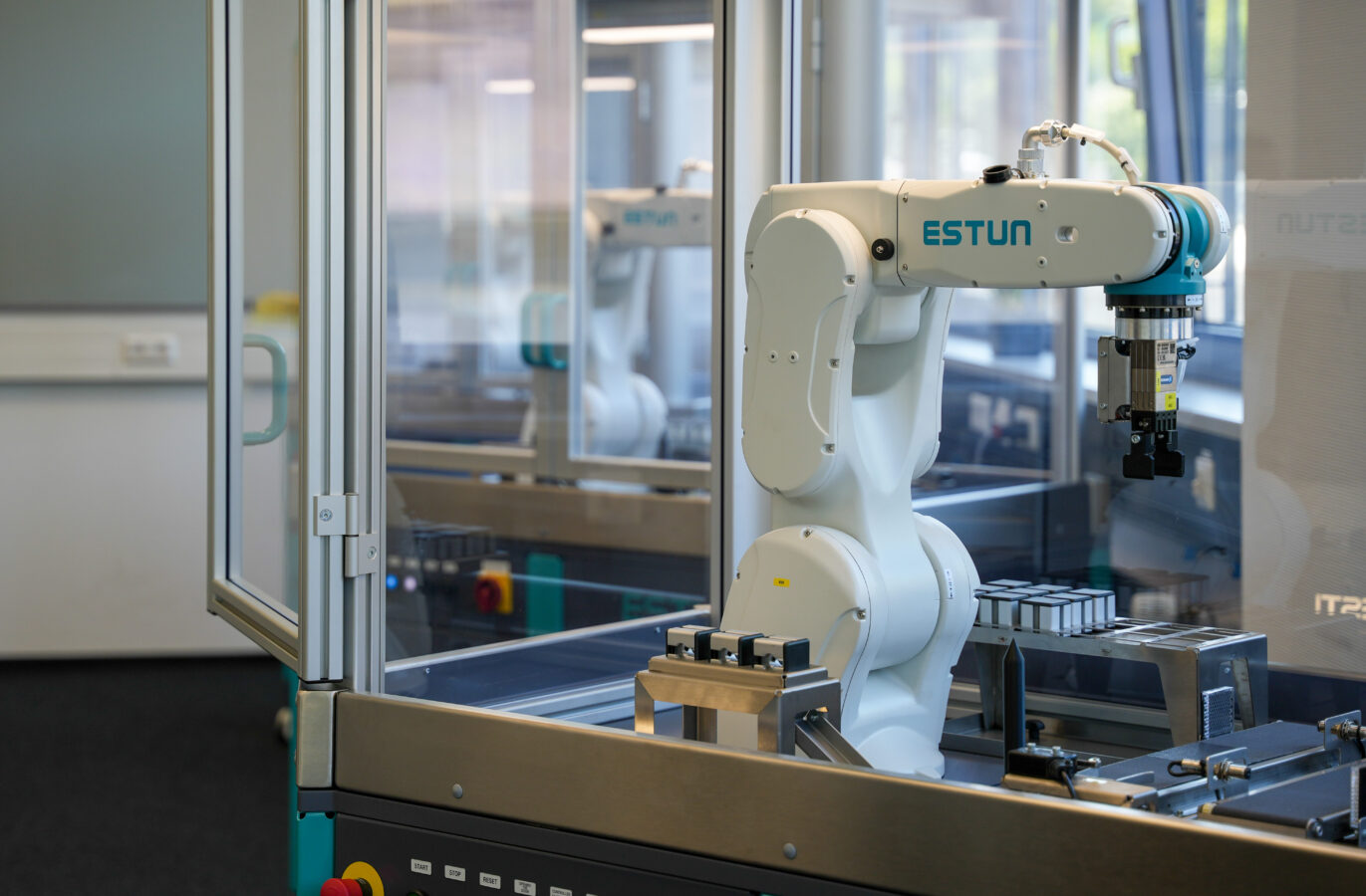 Ein Roboterarm mit der Bezeichnung ESTUN arbeitet in einem verglasten Arbeitsbereich und handhabt Metallteile in einer Industrie- oder Laborumgebung, wobei die Bedientasten und die Ausrüstung in der Nähe sichtbar sind.
