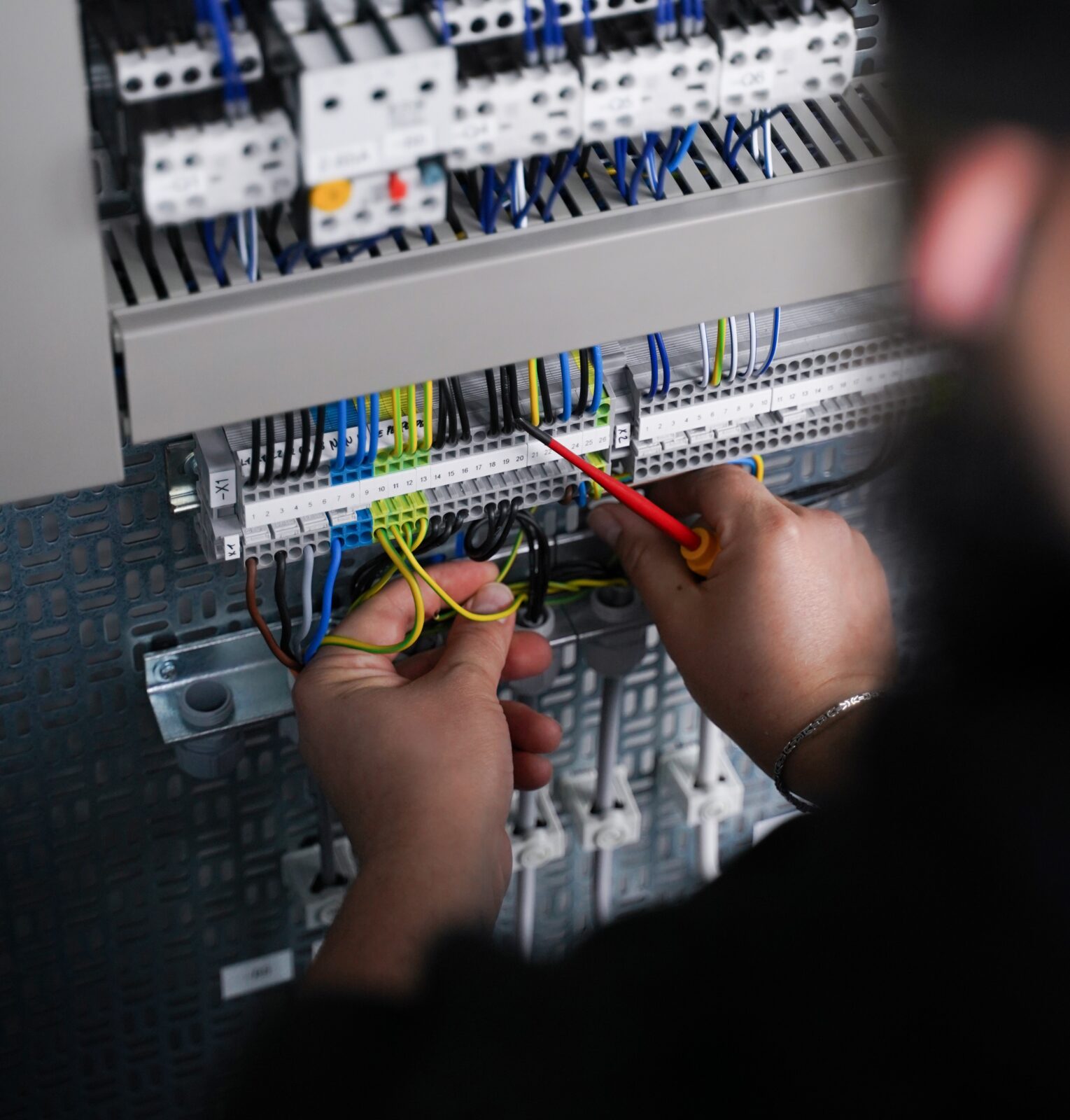 Eine Person arbeitet mit einem roten Schraubendreher an den elektrischen Leitungen in einem Schaltschrank und passt die Verbindungen zwischen den verschiedenfarbigen Kabeln und Klemmen an.