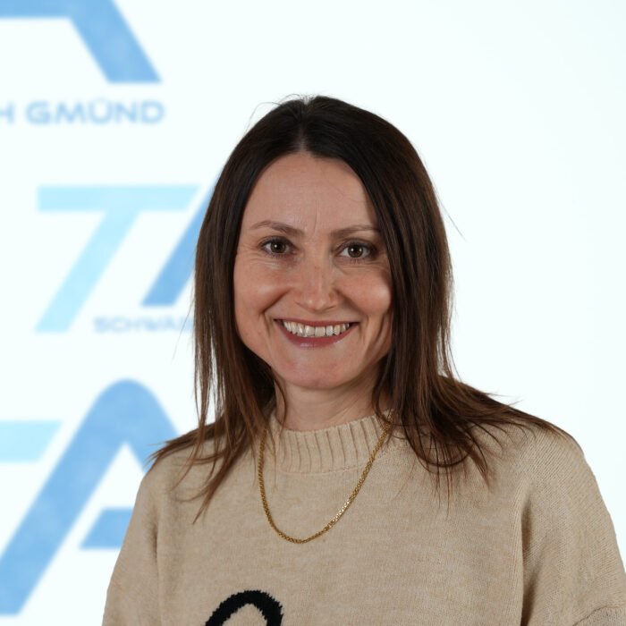 Eine Frau mit glattem braunem Haar, die einen beigen Pullover und eine goldene Halskette trägt, lächelt vor einem weißen Hintergrund mit blauem Text und Logos.