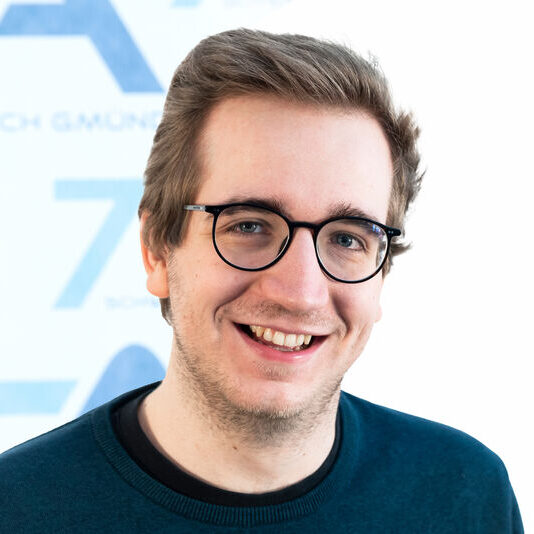 Ein junger Mann mit kurzen hellbraunen Haaren und Brille lächelt in die Kamera; er trägt einen dunkelblaugrünen Pullover. Der Hintergrund ist hell und leicht unscharf, mit blauem Text und Formen.