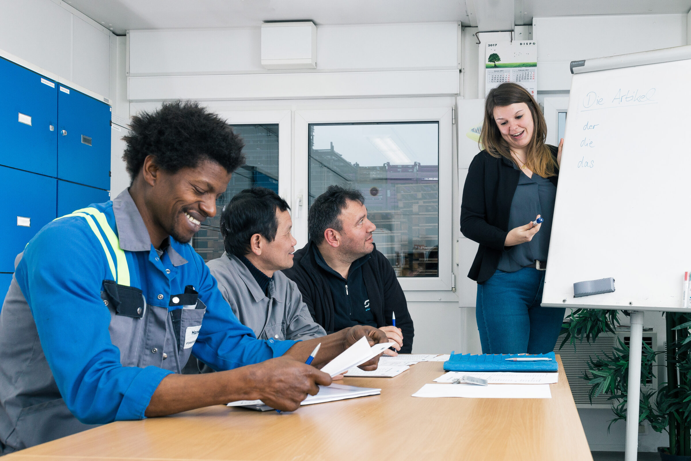 Vier Erwachsene sitzen an einem Tisch und machen sich Notizen, während eine Frau vor einer Weißwandtafel steht und den Unterricht leitet. Die Atmosphäre lässt auf ein Klassenzimmer oder eine Schulung schließen.