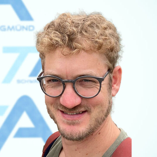 Ein Mann mit lockigem blondem Haar, einem kurzen Bart und einer Brille lächelt in die Kamera. Er trägt ein beiges Hemd und hat einen Taschenriemen über die Schulter geworfen. Im Hintergrund ist ein verschwommenes blau-weißes Schild zu sehen.