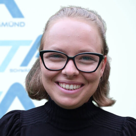 Eine Person mit heller Haut, kurzen blonden Haaren und einer schwarzen Brille lächelt. Sie trägt ein schwarzes Oberteil mit hohem Kragen. Der Hintergrund ist hell und mit blauem Text und Formen versehen, die leicht unscharf sind.
