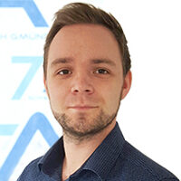 Ein Mann mit kurzen hellbraunen Haaren und einem gepflegten Bart, der ein dunkelblaues Hemd mit Kragen trägt, steht vor einem weißen Hintergrund mit hellblauen geometrischen Formen.