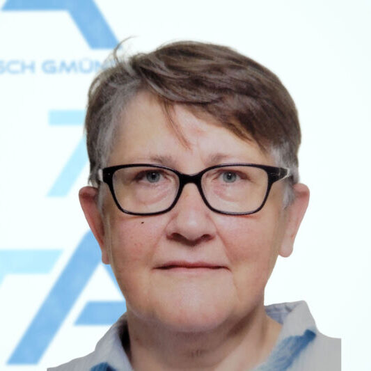 Eine ältere Person mit kurzen grauen Haaren und einer schwarzen Brille blickt in die Kamera; sie trägt ein helles Hemd mit Kragen, im Hintergrund ist ein verschwommenes blau-weißes Bild zu sehen.