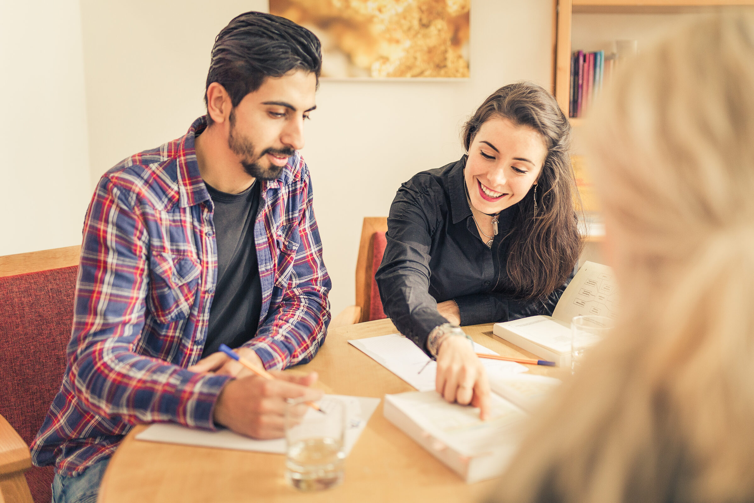 Zwei Personen sitzen mit Notizbüchern und Papieren an einem Tisch und unterhalten sich oder lernen gemeinsam. Eine Person zeigt lächelnd auf eine Seite. Die andere hört aufmerksam zu. Im Vordergrund ist eine dritte Person zu sehen, die unscharf abgebildet ist.