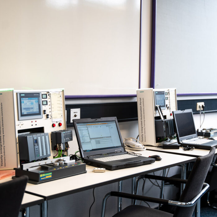 Eine Reihe von Schreibtischen mit Laptops, Bedienfeldern und elektronischen Geräten in einem Klassenzimmer oder Labor. Vor den Schreibtischen stehen schwarze Stühle, und an der Wand im Hintergrund befindet sich eine große Whiteboard-Tafel.