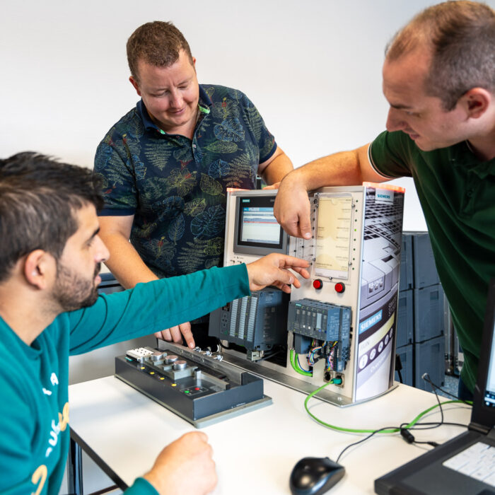 Drei Männer arbeiten gemeinsam an einem Tisch und betrachten ein elektronisches Bedienfeld mit verschiedenen Tasten, Kabeln und einem Bildschirm, auf das sie zeigen. In der Nähe ist außerdem ein Laptop zu sehen.