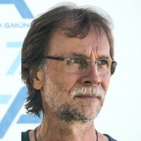 Ein Mann mittleren Alters mit Brille, kurzen braunen Haaren und Bart blickt zur Seite. Er trägt ein dunkles Hemd und steht vor einem hellen Hintergrund mit blauen geometrischen Formen.