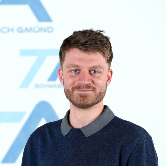 Ein Mann mit kurzen braunen Haaren und Bart, der ein blaues Hemd mit Kragen trägt, lächelt in die Kamera. Der Hintergrund ist weiß und mit blauem Text und Formen versehen, die leicht unscharf sind.