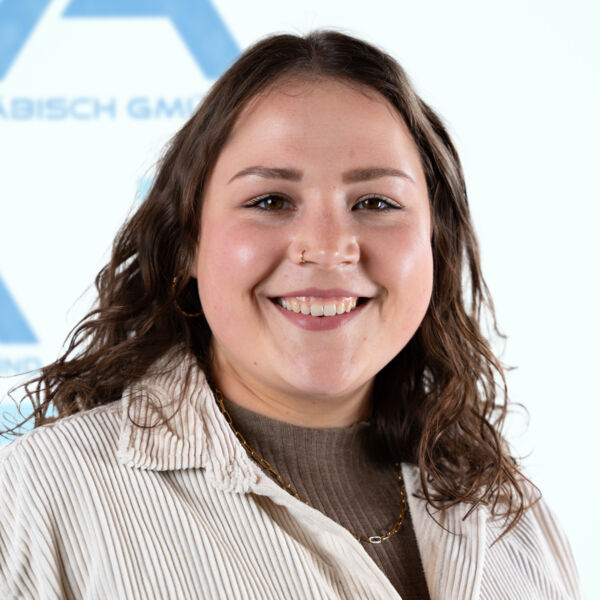 Eine junge Frau mit welligem braunem Haar, die eine beige Jacke über einem braunen Oberteil trägt, lächelt vor einem unscharfen, hellblauen Hintergrund mit Textfragmenten und einem Logo in die Kamera.