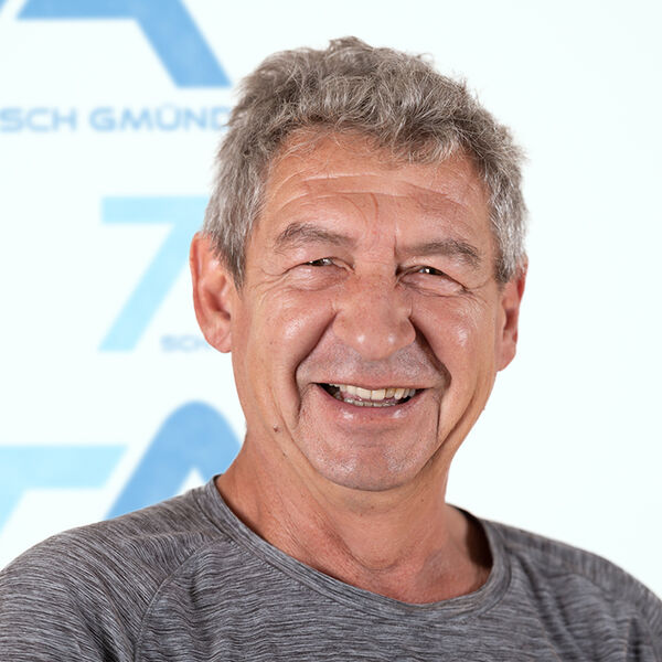 Ein älterer Mann mit grauen Haaren lächelt warmherzig in die Kamera. Er trägt ein graues Hemd und steht vor einem unscharfen weiß-blauen Hintergrund, auf dem teilweise Text und Logos zu erkennen sind.