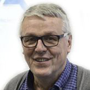 Ein älterer Mann mit kurzen grauen Haaren und Brille, lächelnd, bekleidet mit einem blau karierten Hemd und einem grauen Pullover, der vor einem schlichten hellen Hintergrund steht.