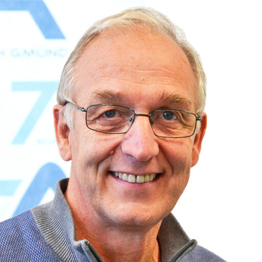 Ein älterer Mann mit kurzen grauen Haaren und Brille lächelt in die Kamera. Er trägt einen blauen Pullover und steht vor einem weißen Hintergrund mit blauem Text und Grafiken.