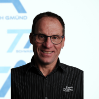 Ein Mann mit kurzen braunen Haaren und Brille, der ein dunkel gestreiftes Hemd mit einem Logo trägt, lächelt vor einem weiß-blauen Hintergrund mit Textfragmenten und geometrischen Formen.