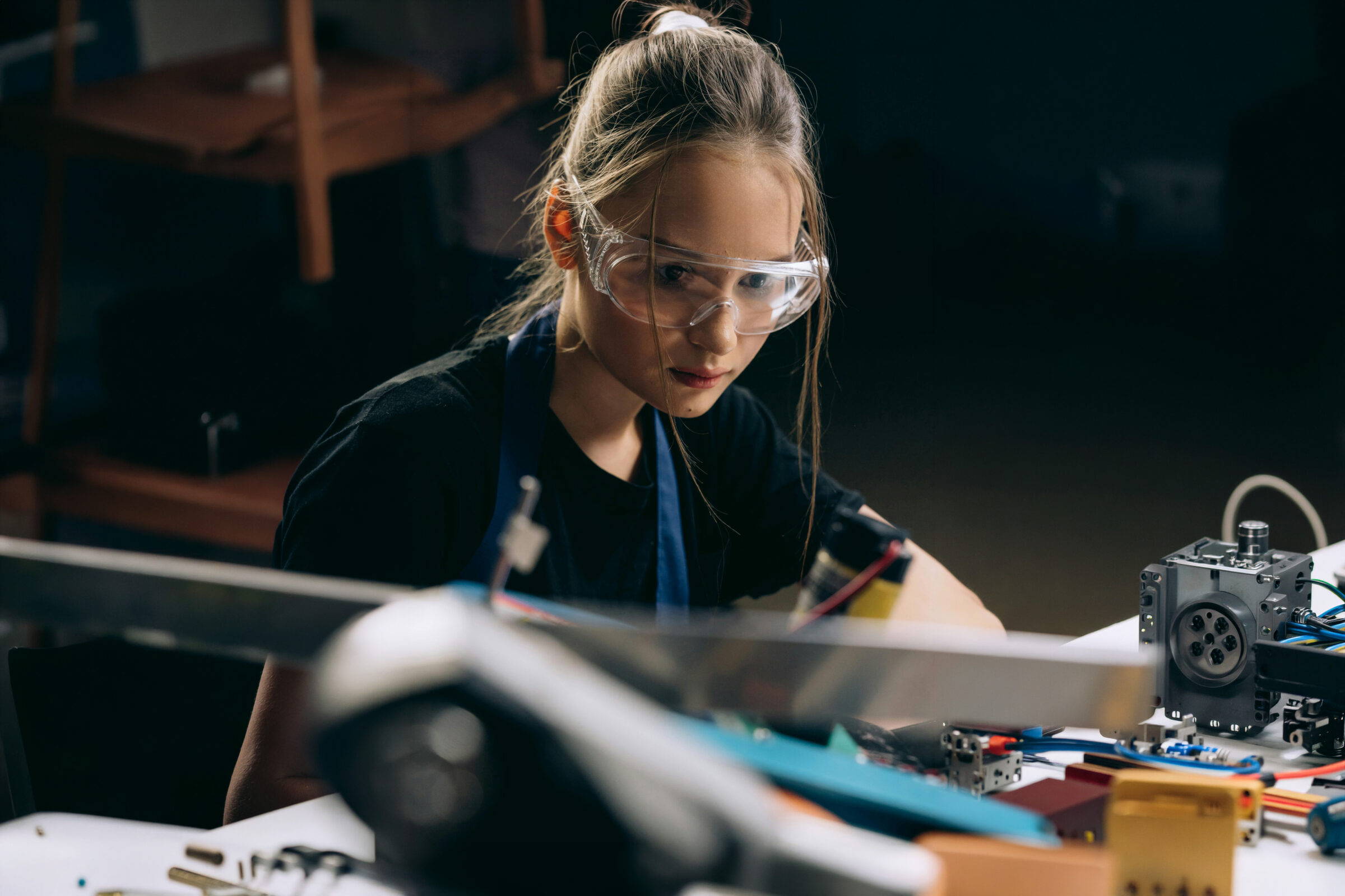 Ein junger Mensch mit Schutzbrille arbeitet konzentriert an einem Robotikprojekt an einem Schreibtisch, der mit elektronischen Bauteilen und Werkzeugen übersät ist.