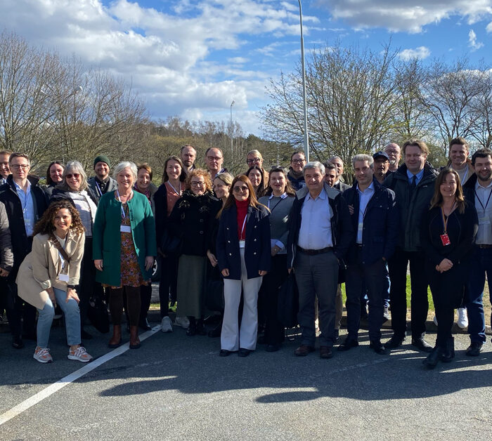 A group of approximately 35 adults, both men and women, pose outdoors on a sunny day against a backdrop of trees and a blue sky. Most are dressed in business or smart-casual attire.