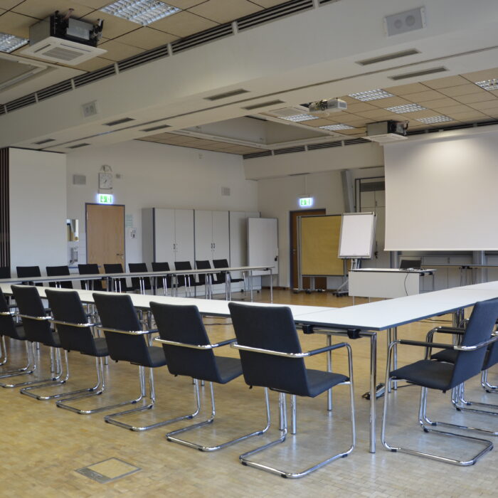 Ein großer Konferenzraum mit U-förmiger Bestuhlung, einer Projektionswand und einem Whiteboard.