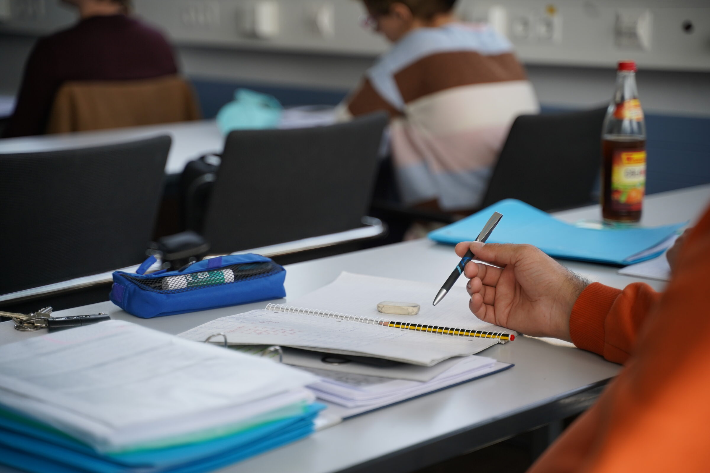 Ein Schüler, der einen Stift über einem Heft auf einem Schreibtisch in einem Klassenzimmer hält.