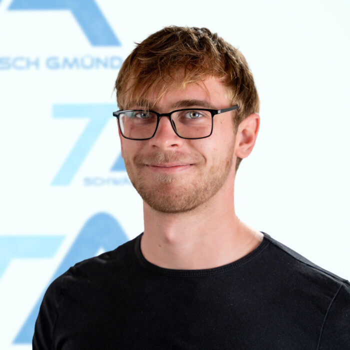 Ein junger Mann mit kurzen braunen Haaren und Brille, der in die Kamera lächelt.