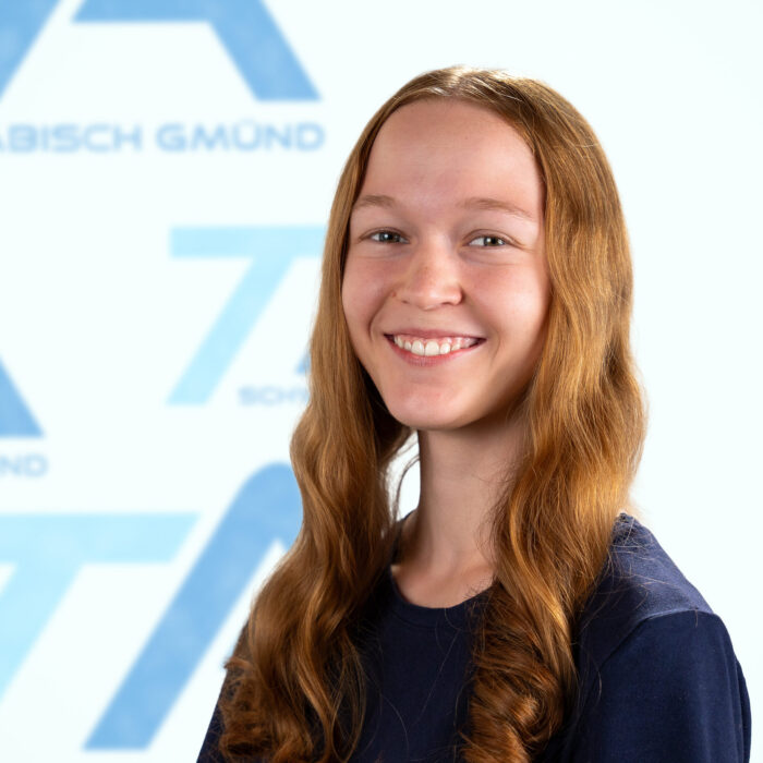 Eine lächelnde junge Frau mit langen rotbraunen Haaren steht vor einem blau-weißen Hintergrund mit Markenlogo.