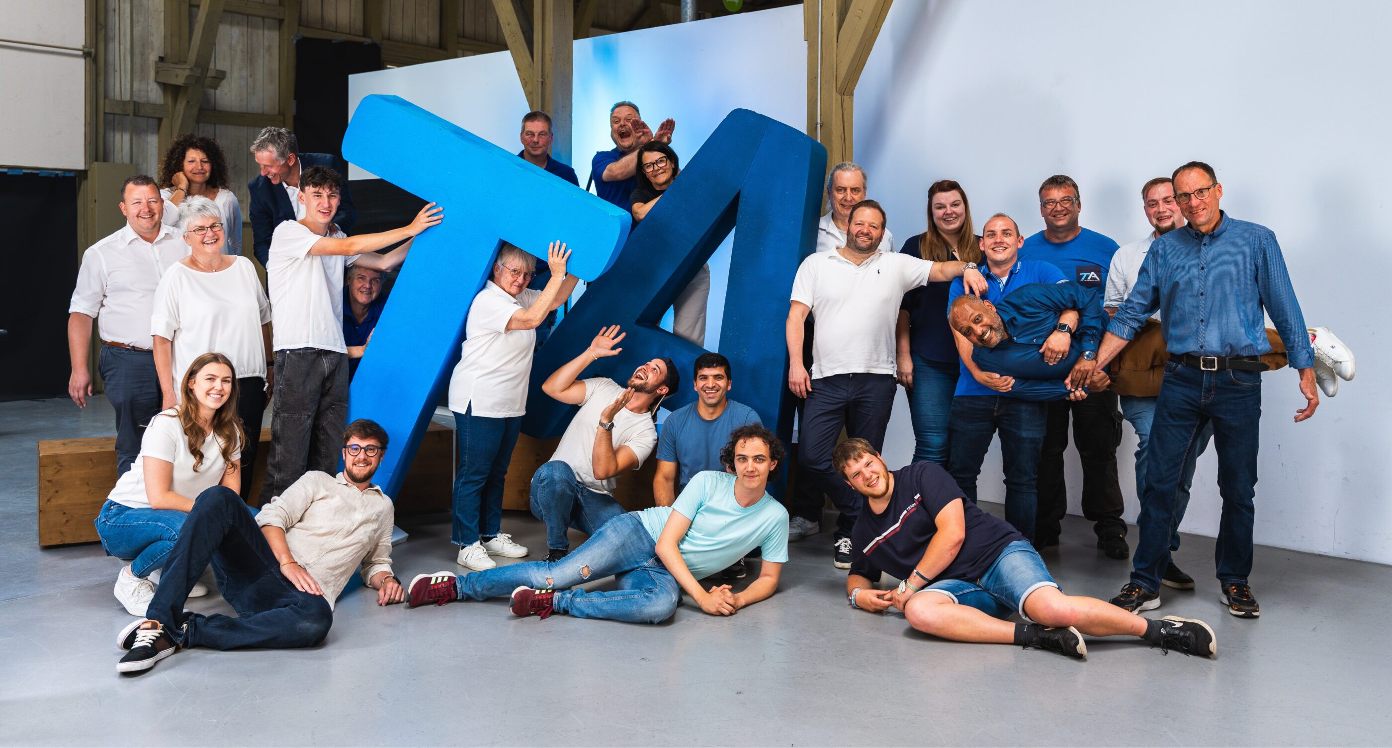 A large group of cheerful people posing, laughing, and playfully gathered around giant, blue 3D letters forming 