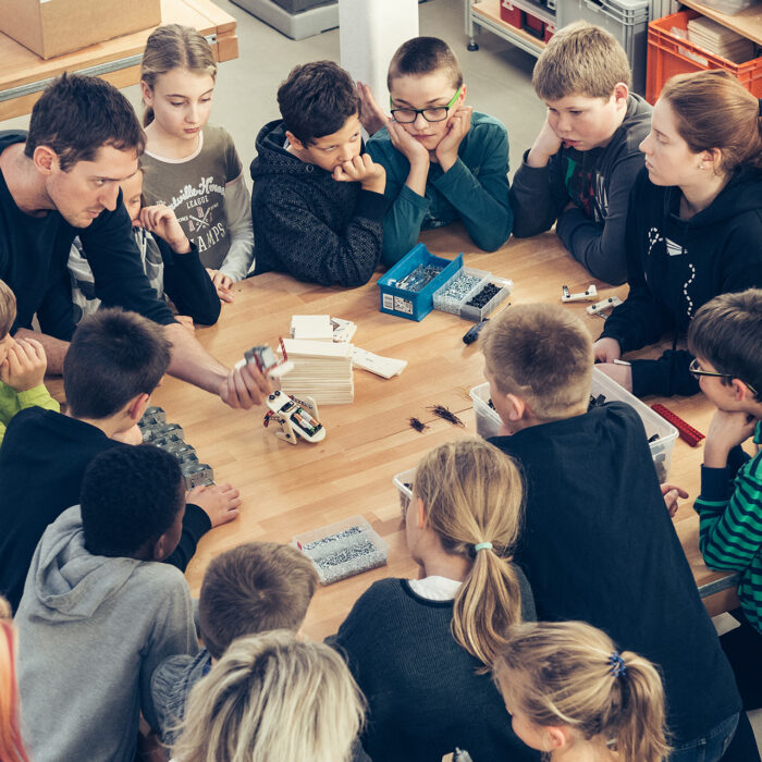 Ein erwachsener Kursleiter demonstriert einer großen Gruppe von Schülern, die sich um einen Holztisch versammelt haben, ein kleines Robotikprojekt.
